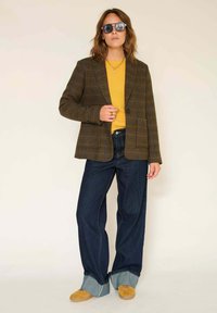 Femme portant des lunettes de soleil noires, un blazer à carreaux marron, un pull jaune, un jean large avec ourlet, et des chaussures jaunes, se tenant devant un fond uni.