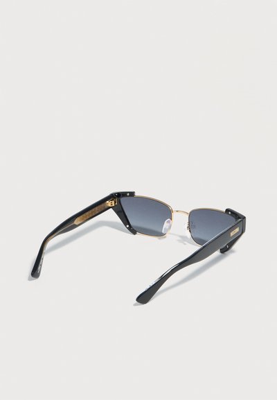 Μαύρα και χρυσά γυαλιά ηλίου με ημίκυρη μεταλλική σκελετό, σκούρα φακούς, διπλωμένα μπράτσα και λογότυπο Moschino στον βραχίονα, εμφανίζονται σε λευκό φόντο.