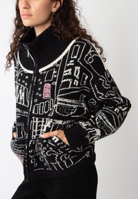 Gebreide zwart-wit cardigan met hoge kraag, voorzien van een grafisch patroon, ritsluiting en voorzakken. Geaccentueerd met roze details.