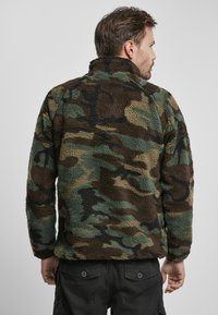 Chaqueta de forro polar con un patrón de camuflaje en verde, marrón y negro, con un cuello alto y una superficie texturizada. Costuras visibles y puños elásticos.
