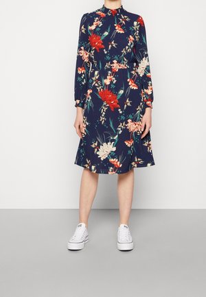 Vrouw draagt een knielange marineblauwe jurk met rode en witte bloemenpatronen en witte sneakers, staand tegen een effen achtergrond.