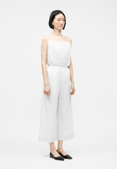 Γυναίκα με κοντά μαύρα μαλλιά που φοράει λευκή ολόσωμη φόρμα χωρίς μανίκια με λεπτές ρίγες και μαύρες μυτερές μπαλαρίνες, στέκεται μπροστά σε απλό φόντο.