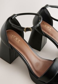 Czarne sandały na blokowanym obcasie z szerokim paskiem z przodu, paskiem wokół kostki i złotą klamrą. Posiadają gładką nude wkładkę z logo marki.