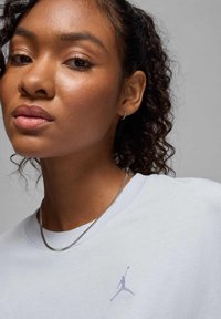 T-shirt azzurro chiaro con una texture morbida, caratterizzato da un logo Jumpman ricamato bianco sul lato sinistro del petto. Accento con collana metallica sottile.