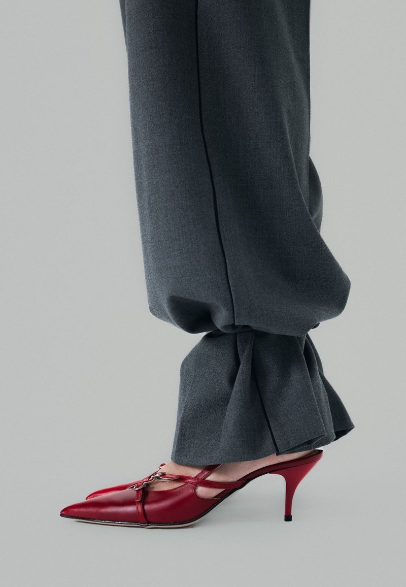 Rode leren pumps met een spits toelopende neus, een slingback ontwerp en een kleine hak, gecombineerd met grijze stoffen broeken met een knoopdetail aan de zoom.