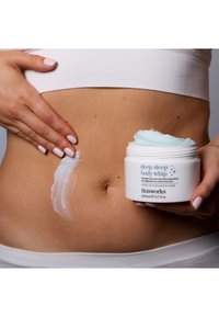Deep Sleep Body Whip i en hvit krukke med blå detaljer, kremaktig tekstur påført huden, ved siden av en hånd som forsiktig jevner ut kremen.