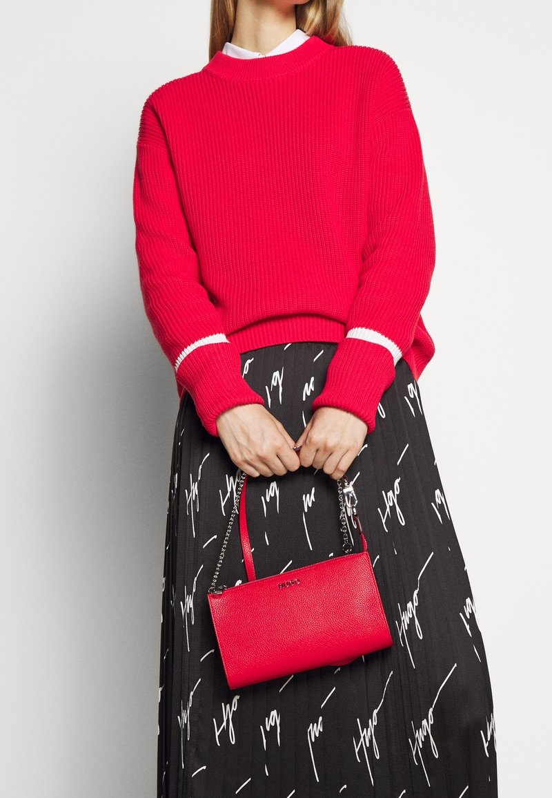 Femme portant un pull rouge vif et une jupe noire avec une inscription blanche, tenant un petit sac à main rouge avec une bandoulière chaîne devant un fond blanc.