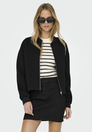 Chaqueta bomber corta negra con cierre de cremallera, combinada con un suéter a rayas en blanco y azul marino y una falda de mezclilla negra de cintura alta.