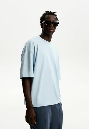 Light blue oversized t-shirt made of soft cotton, featuring a round neckline and short sleeves, worn with dark trousers and sunglasses.