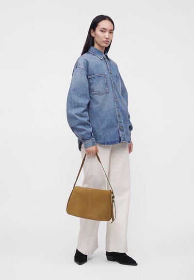 Femme portant une chemise en jean bleue oversize, pantalon blanc, chaussures noires, tenant un sac à main en daim marron à ses côtés sur un fond uni.