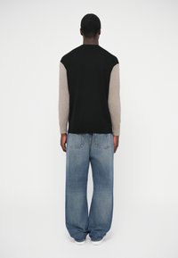 Πουλόβερ με χρωματικό μπλοκ, μαύρο σώμα και ανοιχτό καφέ μανίκια, συνδυασμένο με φαρδιά τζιν μπλε, από πίσω, σε απλό φόντο.