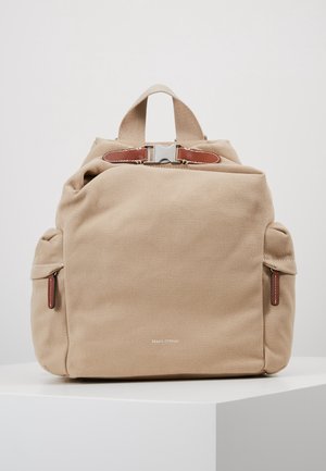 Beigefarbener Stoffrucksack mit zwei Seitentaschen, einem oberen Tragegriff, braunem Lederriemen mit silberner Schnalle und minimalem Frontlogo auf weißer Oberfläche.