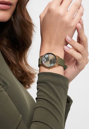 Die Uhr verfügt über ein Gehäuse in Goldton mit einem blumengemusterten Ziffernblatt, einem grünen Silikonarmband und minimalistisch gestalteten Stundenmarkierungen.