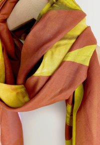 Szalik wykonany z miękkiego materiału, z rdzawymi i jasnożółtymi paskami. Ma gładką teksturę i swobodny, opływowy wygląd.