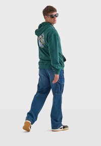 Sudadera verde con capucha y bolsillo tipo canguro en la parte delantera, con un gráfico colorido en la espalda. Combinada con vaqueros cargo azules sueltos y zapatillas.
