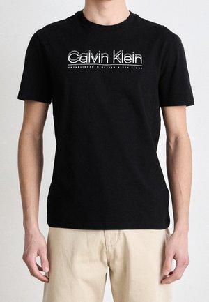 Osoba nosząca czarną krótką koszulkę z napisem „Calvin Klein” i rokiem założenia nadrukowanym białą czcionką na piersi.