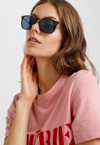Óculos de sol com padrão tartaruga e lentes escuras; forma retangular. Modelo vestindo uma t-shirt rosa com texto vermelho, mão tocando o pescoço.