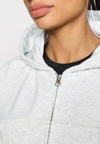 Sudadera con capucha gris claro con cremallera, confeccionada en un tejido suave y texturizado, con un bolsillo frontal y una cremallera plateada, combinada con una camiseta negra.