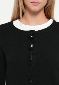 Junge Frau, die eine schwarze Knopfjacke über einem weißen Rundhalsausschnitt trägt, von den Schultern bis zu den Lippen vor einem weißen Hintergrund abgebildet.