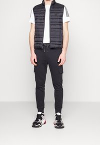 Mężczyzna w czarnej zapinanej kamizelce, białej koszuli z krótkim rękawem i paskami na rękawach, czarnych dżoggerach typu cargo oraz czarno-białych sneakersach.