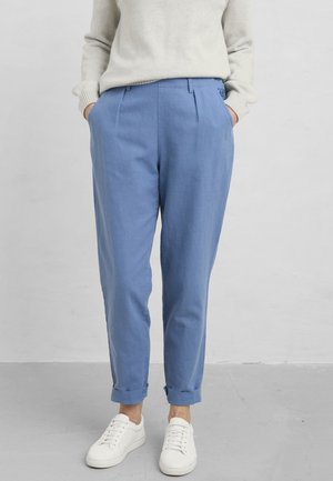 Person wearing light blue tailored pants with cuffs, cream sweater, white sneakers, standing against a plain light gray background.