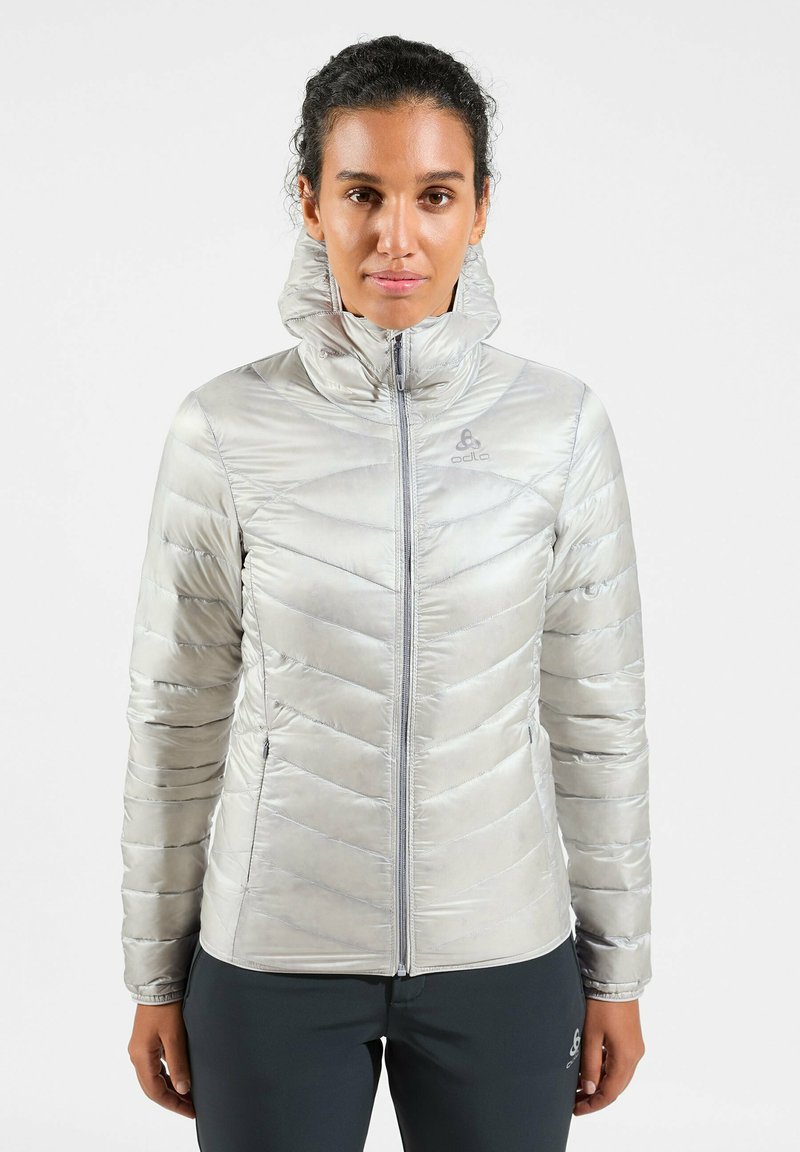 Helle silberne isolierte Jacke mit einem Zickzackmuster, hohem Kragen, vorderem Reißverschluss und Logo auf der Brust. Glatte, glänzende Stofftextur.