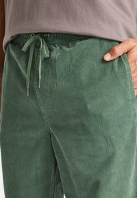 Green corduroy pants with a textured finish, featuring an elastic waistband and drawstring closure. Side pockets visible.