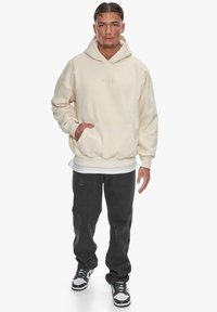Beige Kapuzenpullover mit einer Fronttasche und gerippten Bündchen, über einem weißen Shirt getragen. Dunkelgraue Jogginghosen und schwarz-weiße Sneaker runden den Look ab.
