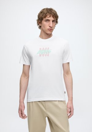 Jeune homme aux cheveux bouclés portant un T-shirt blanc avec un logo « BOSS » pastel et un pantalon beige sur un fond blanc uni.