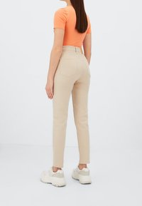 Vaqueros beige de cintura alta con un diseño ajustado, que cuentan con dos bolsillos traseros y una etiqueta en la cinturilla. Combinados con una blusa de manga corta naranja y zapatillas blancas.