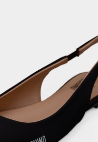 Κοντινή λήψη μαύρου επίπεδου παπουτσιού τύπου slingback με ταμπά εσωτερική επένδυση και καμπυλωτό λουρί πάνω από τη φτέρνα σε λευκό φόντο.