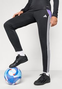 Černé sportovní kalhoty s bílými a fialovými bočními akcenty, vybavené třemi vertikálními bílými pruhy. Model stojí na modro-červeném fotbale.
