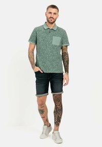 Groene gepatternde poloshirt met een zak, gecombineerd met donkere denim shorts en witte sneakers. Zichtbare tatoeages op armen en benen.