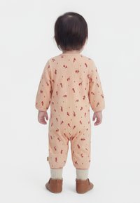 Maluszek stojący tyłem, ubrany w brzoskwiniowy kombinezon w kwiatowy wzór, kremowe skarpetki i brązowe buty na białym tle.