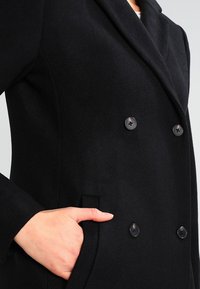 Mustat villasta valmistettu kaksinkertainen takki, jossa on leveä kaulus, kaksi nappia, sivutaskut ja muotoiltu istuvuus. Suunnikas pinta.