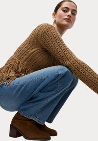 Stickad tröja i brunt med hålmönster och fransdetalj, matchad med ljusblå vida jeans och mörkbruna ankelstövlar.