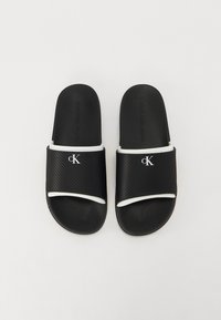 Musta kumisaapassandaalit, joissa on teksturoitu hihna, valkoiset yksityiskohdat ja hopea "cK" -logo päällä. Litteä pohja, joka on suunniteltu mukavuuteen.