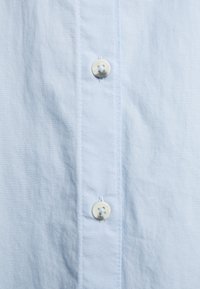 Ljust blått tyg med två vita sydda knappar ordnade vertikalt på en skjortslå med synlig söm.