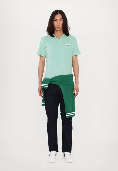 Lys grønn poloskjorte med krage, iført en grønn genser knyttet rundt livet, mørke jeans og hvite joggesko, stående mot en hvit bakgrunn.
