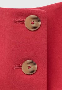 Červená látka s texturovaným vzhledem, která má dva velké, kulaté, lesklé knoflíky. Každý knoflík má uprostřed malý vyšitý detail.