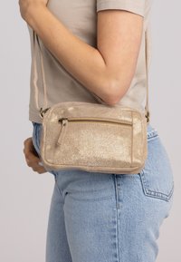 Donna che indossa jeans azzurri chiari e una maglietta beige, mentre porta una piccola borsa a tracolla dorata luccicante con una tasca con zip frontale.