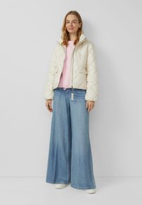 Kremowa pikowana kurtka z zamkiem z przodu, w parze z różowym T-shirtem i szerokimi dżinsami o luźnym kroju. Sneakersy mają teksturowaną białą podeszwę.