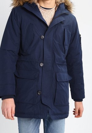 Man draagt een marineblauwe winterparka met bontvoering aan de kap, knopen en rits aan de voorkant, over een beige gebreide trui en blauwe spijkerbroek.