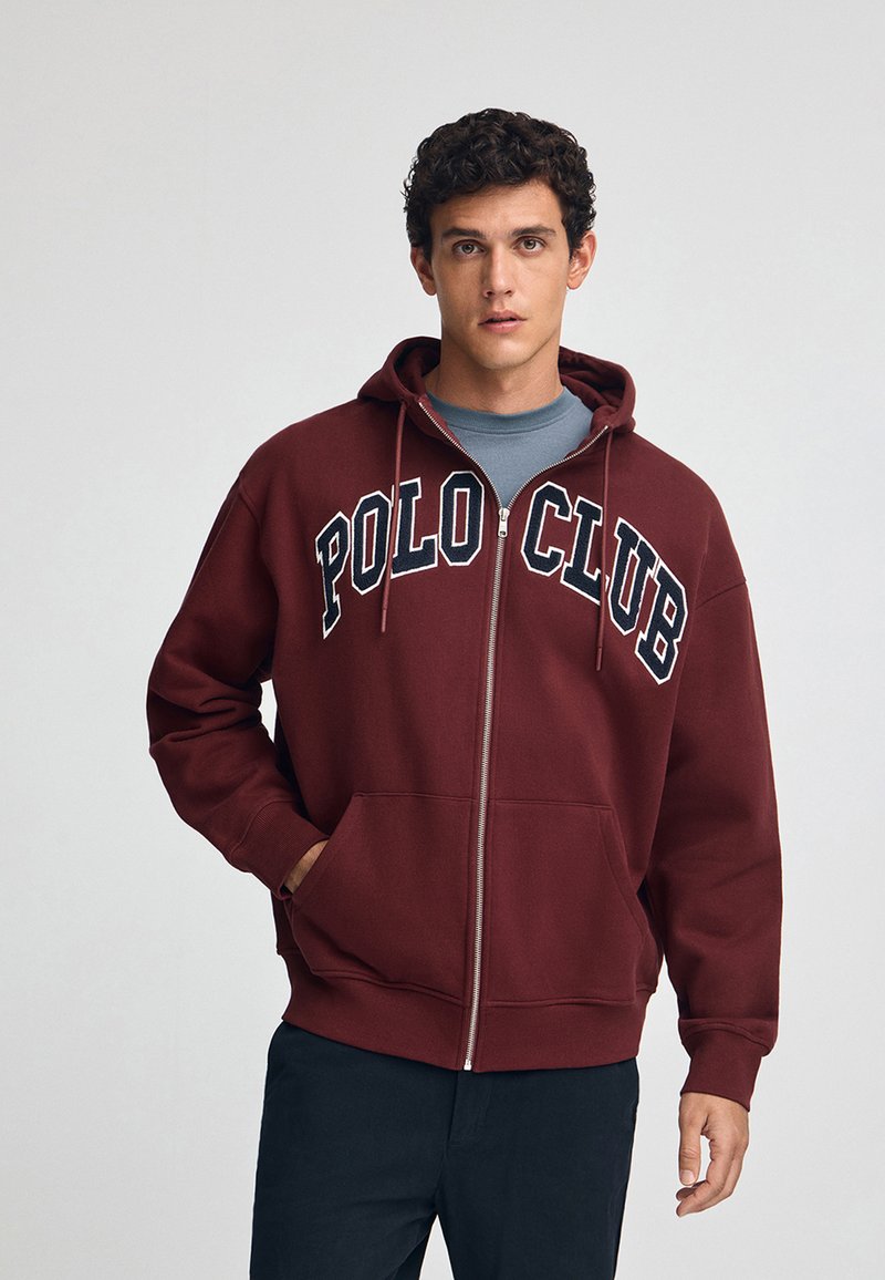 Burgundy zip-hoodie med en framficka i känguru stil och "POLO CLUB"-logotyp i vitt på bröstet. Tyget har en mjuk och strukturerad yta.