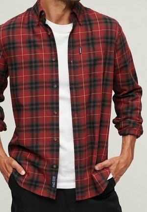 Mand iført en rød og sort ternet skjorte med knapper over en hvid t-shirt med rund hals, med hænderne hvilende i lommerne på sorte bukser.