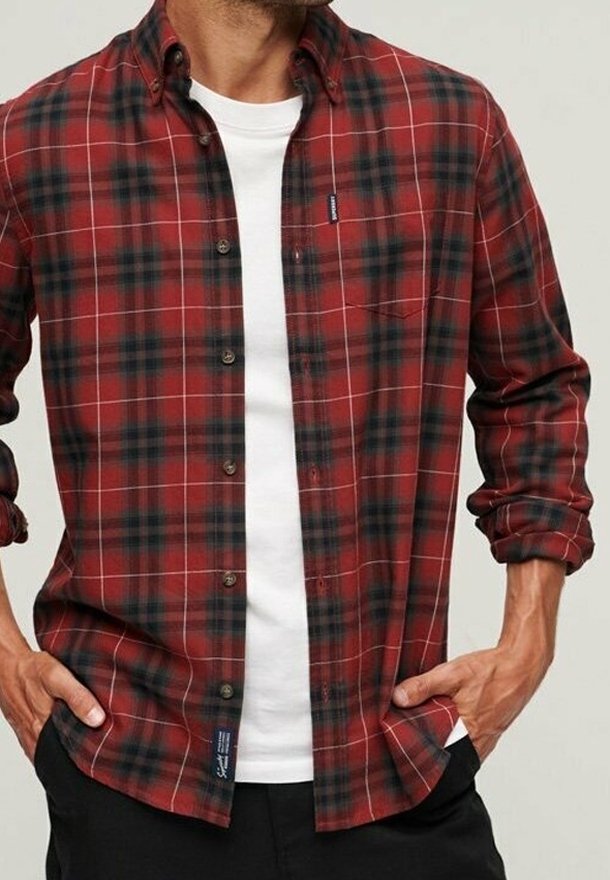 Homme portant une chemise à carreaux rouge et noir boutonnée sur un t-shirt blanc à col rond, les mains dans les poches d'un pantalon noir.