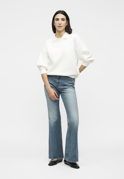 Femme debout portant un sweat-shirt blanc à manches longues, un jean flare bleu et des ballerines noires pointues sur un fond blanc uni.