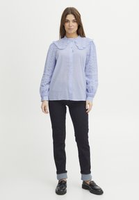 Svetlo modra srajca s gumbki z velikim nabranim ovratnikom in vezenimi čipkastimi rokavi. Združena z temnimi kavbojkami in črnimi loaferji.