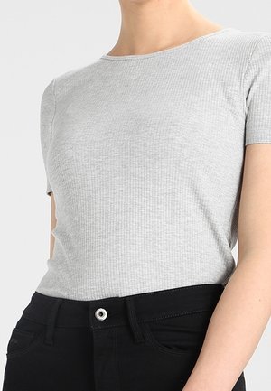 Persoon die een aansluitend lichtgrijs, geribd shirt met korte mouwen draagt, ingestopt in een zwarte broek met hoge taille en knoopsluiting.