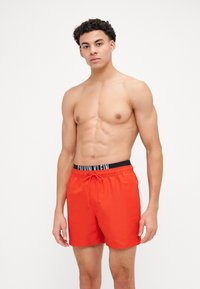 Shorts de bain rouges avec une taille élastique noire arborant le logo "CALVIN KLEIN", fermeture à cordon de serrage et tissu lisse et léger.